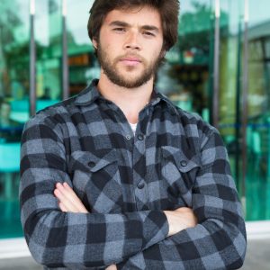 Serious young man standing outdoors with his arms crossed. Handsome guy wearing casual shirt, looking at camera and standing with glass wall in background. Young man portrait concept. Front view.